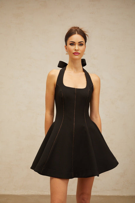 Young woman wearing a black halterneck mini dress with bow detail at the neck standing against a plain background