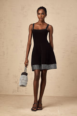 Young woman wearing a black knit mini dress with flared skirt and studded details standing in front of plain background