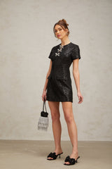 Young woman wearing a black mini dress with glitter details and short sleeves standing against a plain background