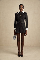 Young woman wearing a black mini dress with sequin panels and tweed fabric standing in front of a plain background