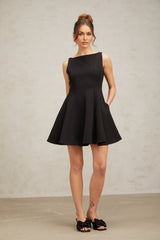 Young woman wearing a black sleeveless mini dress with a flared skirt standing against a plain background