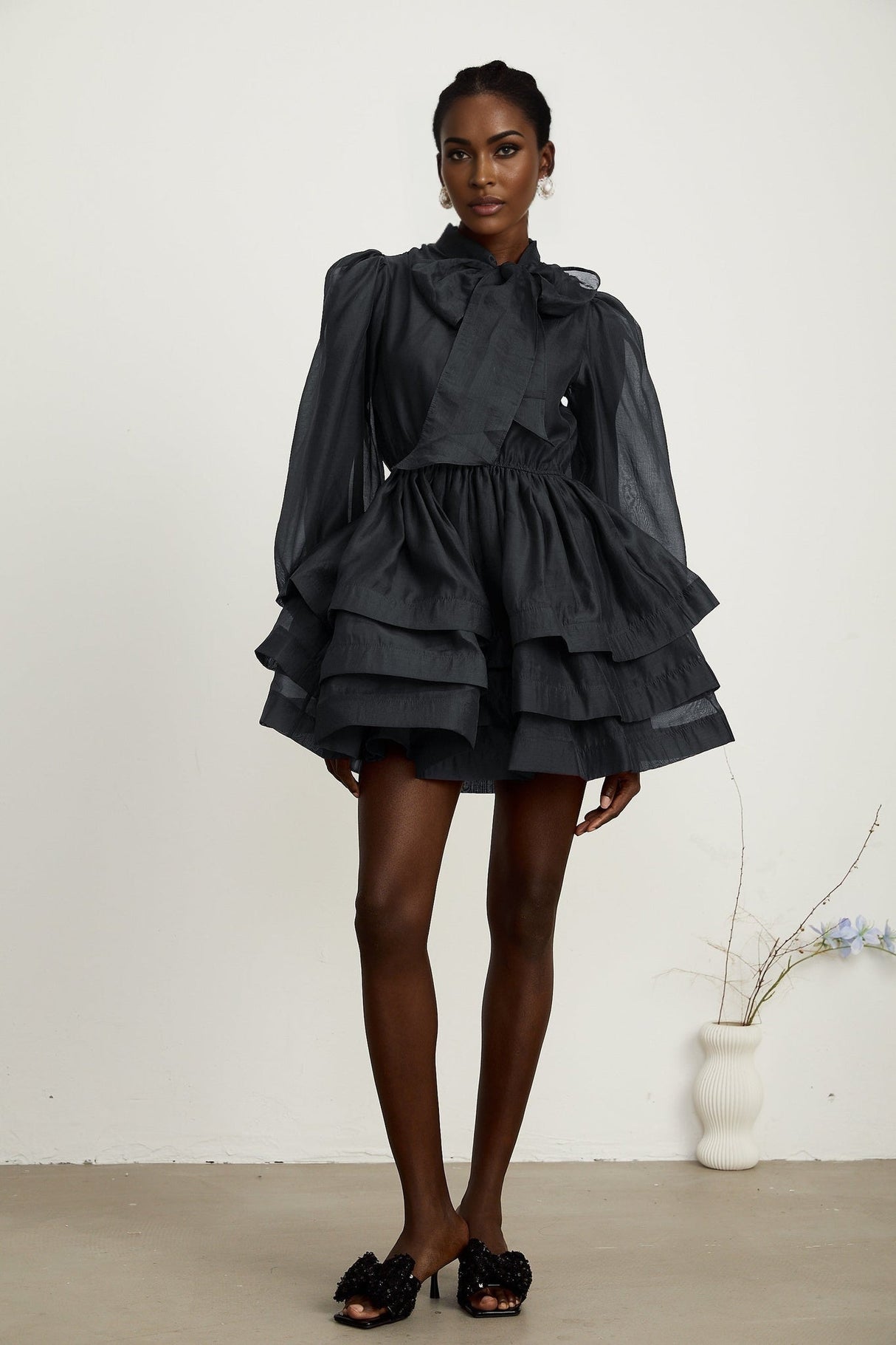 Young woman wearing a black tiered flared mini dress with short sleeves and a vneckline standing against a plain background
