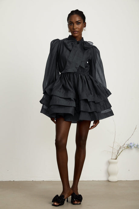 Young woman wearing a black tiered flared mini dress with short sleeves and a vneckline standing against a plain background