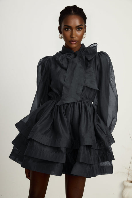 Young woman wearing a black tiered flared mini dress with short sleeves standing against a plain background