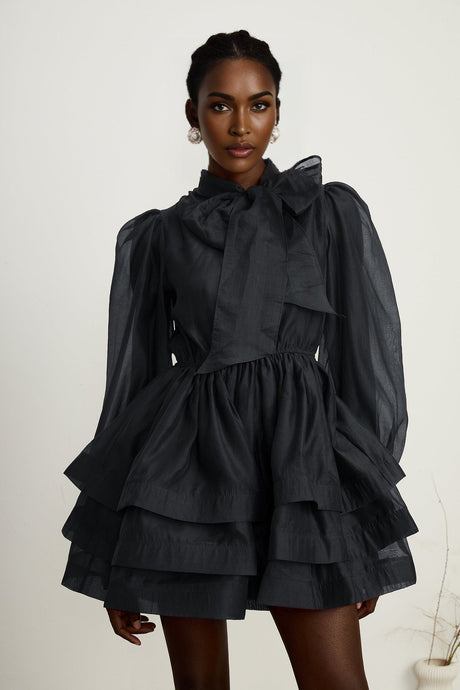 Young woman wearing a black tiered flared mini dress with short sleeves standing against a plain background