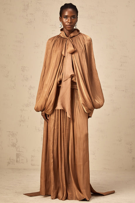 Young woman wearing a long brown pleated satin maxi dress with a flowing skirt and fitted bodice standing in a neutral setting
