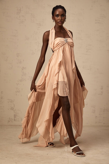 Young woman wearing a long pink chiffon maxi dress with ruffles and halter neck style standing in front of a plain background