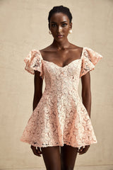 Young woman wearing a pink lace mini dress with flutter sleeves standing against a plain background