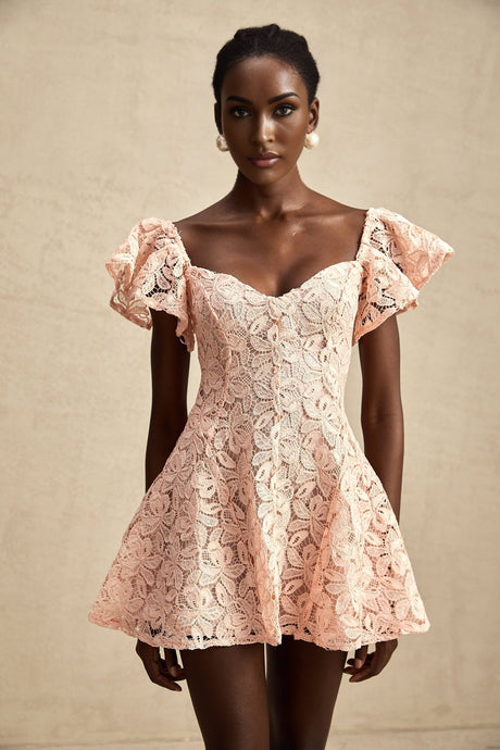 Young woman wearing a pink lace mini dress with flutter sleeves standing against a plain background