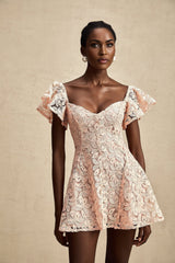 Young woman wearing a pink lace mini dress with flutter sleeves standing against a plain background