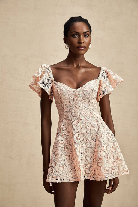 Young woman wearing a pink lace mini dress with flutter sleeves standing against a plain background