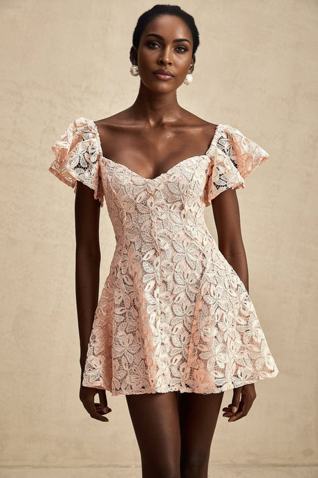 Young woman wearing a pink lace mini dress with flutter sleeves standing in front of a plain background