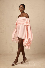Young woman wearing a pink mini dress with bow detail and draped fabric flowing down the front standing against a plain background