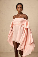 Young woman wearing a pink mini dress with bow detail and draped fabric standing against a plain background
