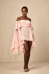 Young woman wearing a pink mini dress with bow detail and draped fabric standing against a plain background