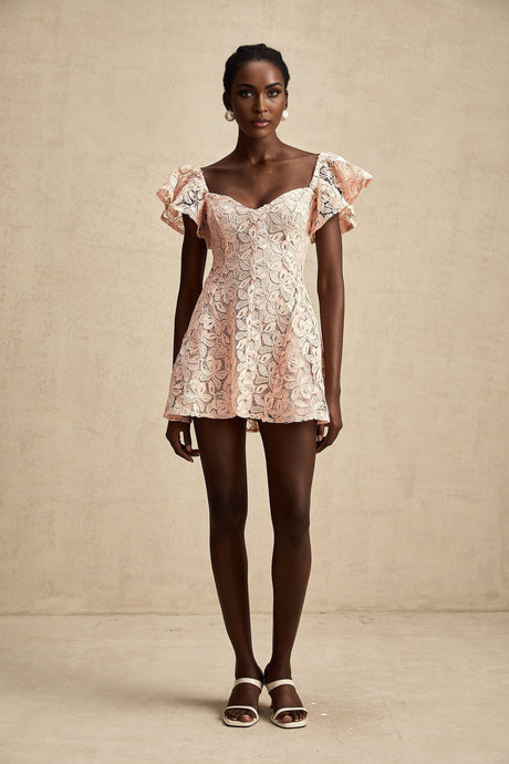 Young woman wearing a pink mini dress with flutter sleeves and lace detailing standing in a relaxed pose against a plain background