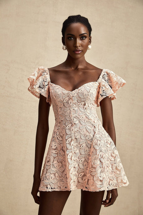 Young woman wearing a pink mini dress with lace details and flutter sleeves posing against a plain background