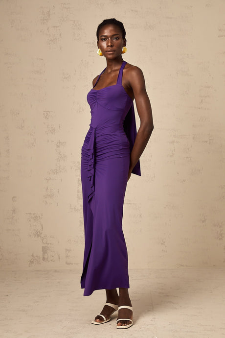 Young woman wearing a purple halterneck midi dress with ruched detailing standing against a plain background