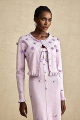 Young woman wearing a purple knitted cardigan and matching dress with faux flower embellishments standing against a plain background