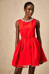 Young woman wearing a red mini dress with pleated skirt and embellished bodice standing in front of plain background