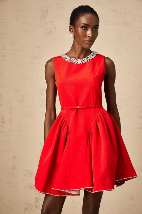 Young woman wearing a red mini dress with pleated skirt and embellished bodice standing in front of plain background
