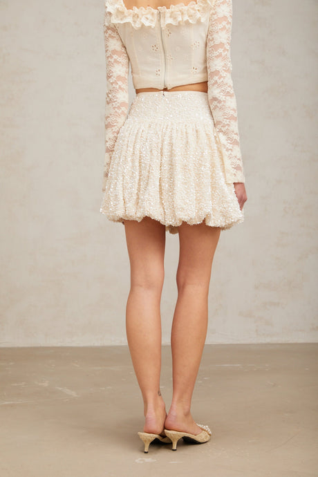 Young woman wearing a shimmering pleated mini skirt with sequin details standing against a plain background