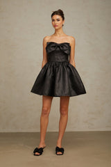 Young woman wearing a short black metallic dress with a bow detail standing in front of a plain background