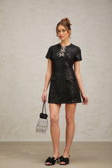 Young woman wearing a short black mini dress with glitter details standing in a neutral pose against a plain background