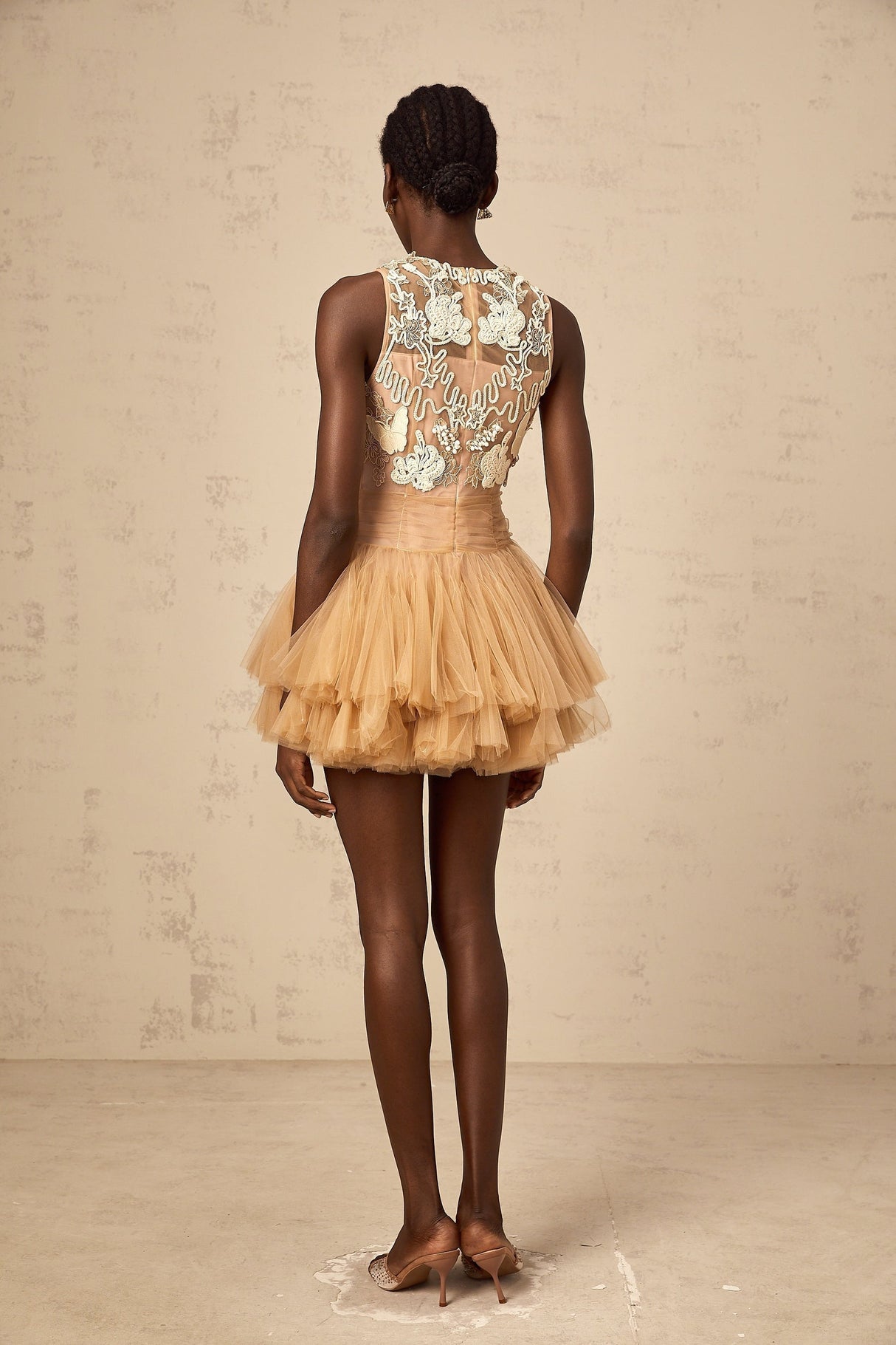 Young woman wearing a short khaki dress with corded lace panels and embellishments standing against a plain background