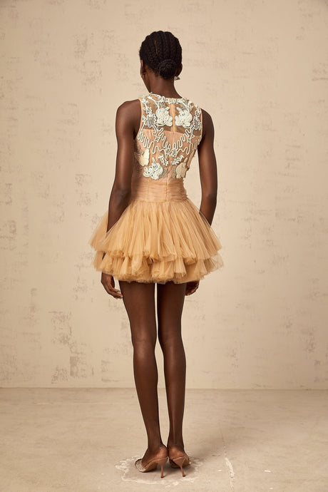 Young woman wearing a short khaki dress with corded lace panels and embellishments standing against a plain background