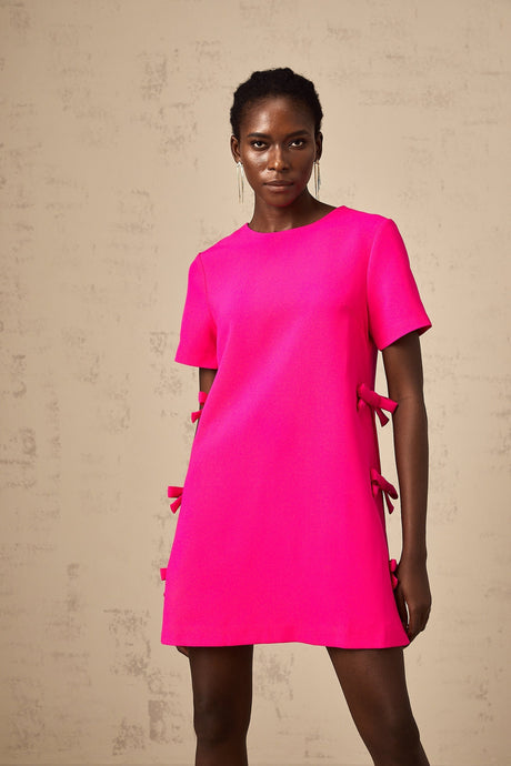 Young woman wearing a short pink dress with bow detail standing in a neutral background