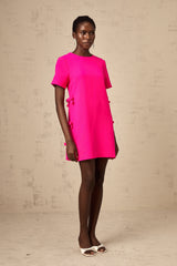 Young woman wearing a short pink dress with bow details on the front standing against a plain background