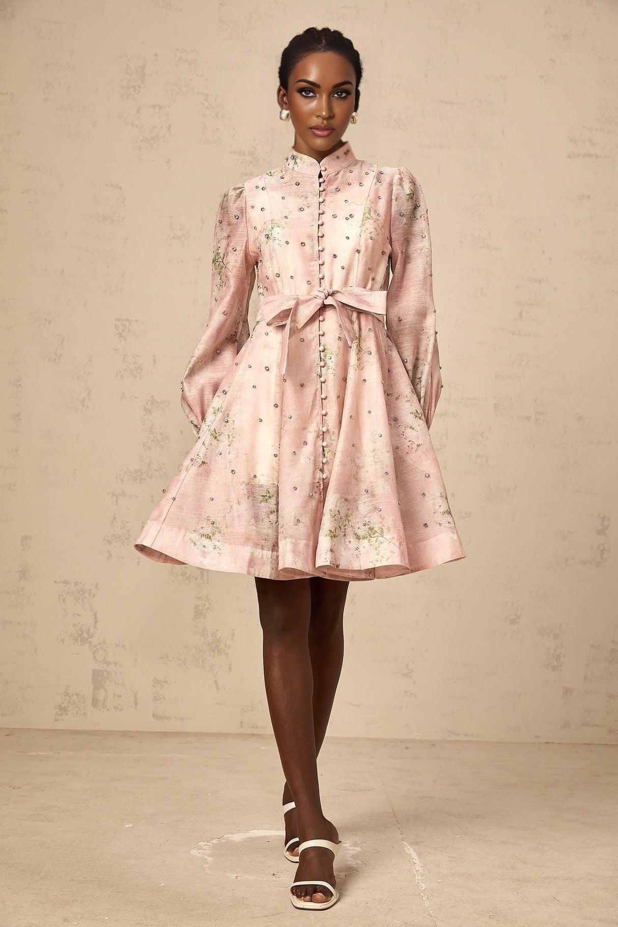 Young woman wearing a short pink dress with floral print and embellishments posing against a plain background
