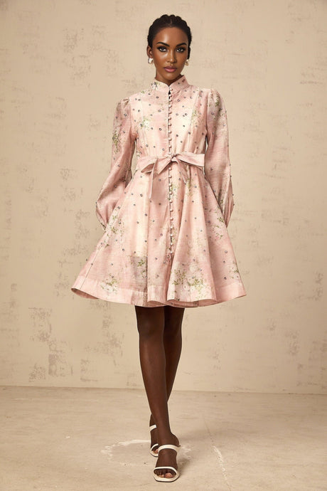 Young woman wearing a short pink dress with floral print and embellishments posing against a plain background
