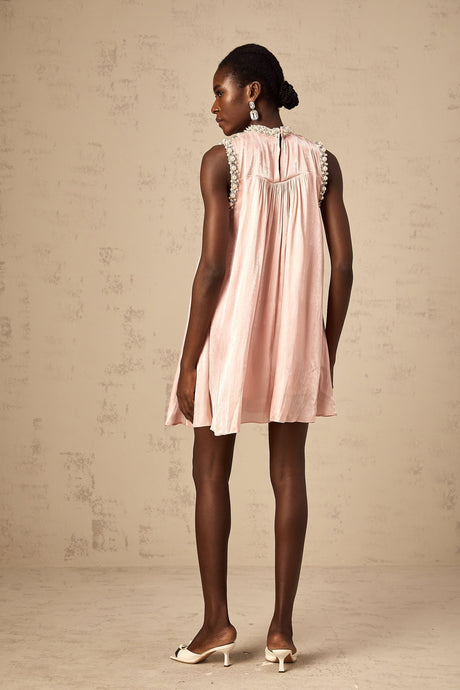 Young woman wearing a short pink dress with pleated skirt and embellished bodice standing against a plain background