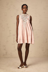 Young woman wearing a short pink dress with pleated skirt and embellished bodice standing in front of plain background