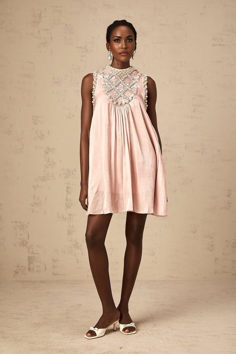 Young woman wearing a short pink dress with pleated skirt and embellished bodice standing in front of plain background