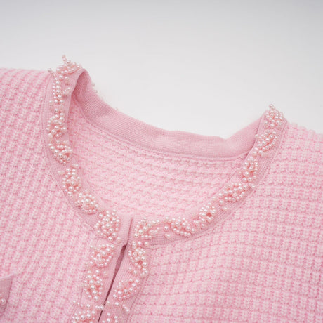 Young woman wearing a short pink knitted dress with beaded embellishments standing against a plain background