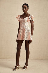Young woman wearing a short pink lace dress with flutter sleeves and a fitted silhouette standing against a plain background