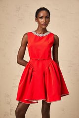 Young woman wearing a short red dress with pleated skirt and embellished details standing against a plain background