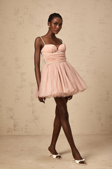 Young woman wearing a short sleeveless dress with a pleated tulle skirt in a light color against a plain background