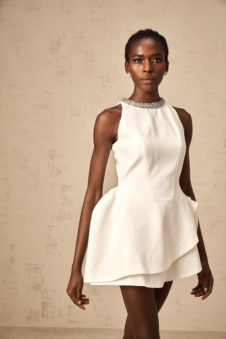Young woman wearing a short white crystalembellished mini dress with layered skirt and spaghetti straps standing against a plain background