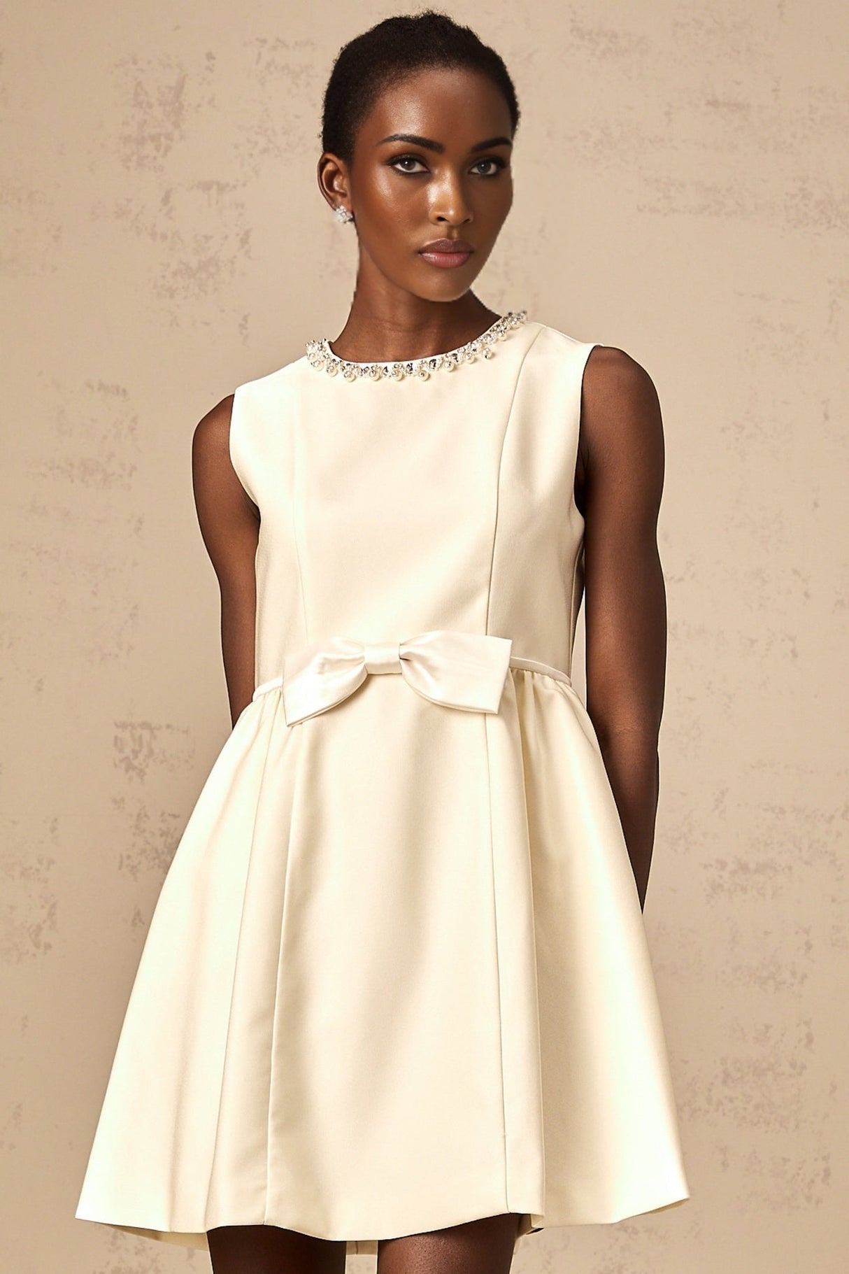 Young woman wearing a short white dress with a bowknot detail and embellishments standing against a plain background