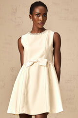 Young woman wearing a short white dress with a bowknot detail and embellishments standing against a plain background