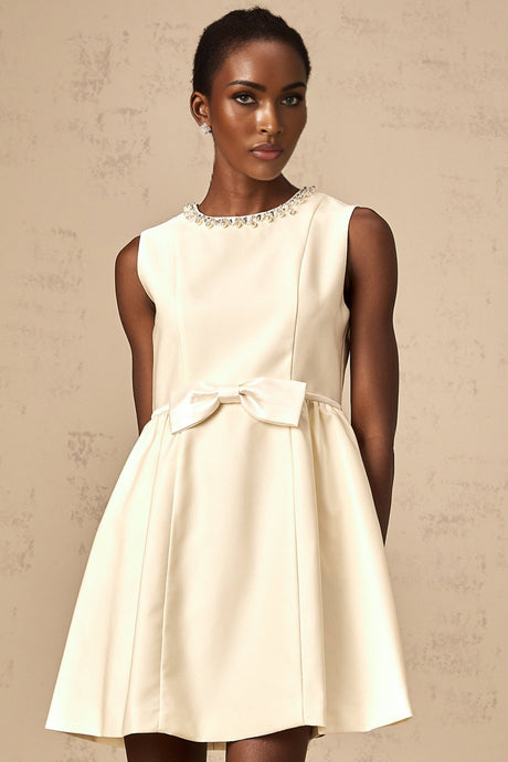 Young woman wearing a short white dress with a bowknot detail and embellishments standing against a plain background