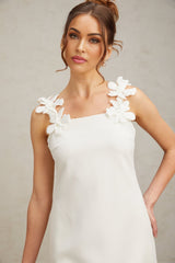 Young woman wearing a short white dress with faux flower details standing in a casual pose against a plain background