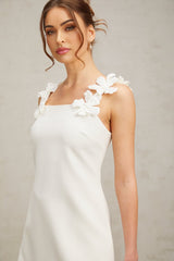 Young woman wearing a short white dress with faux flower embellishments standing against a plain background