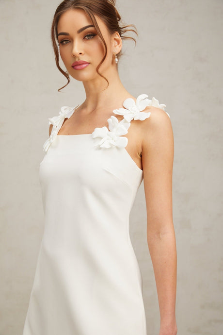 Young woman wearing a short white dress with faux flower embellishments standing against a plain background