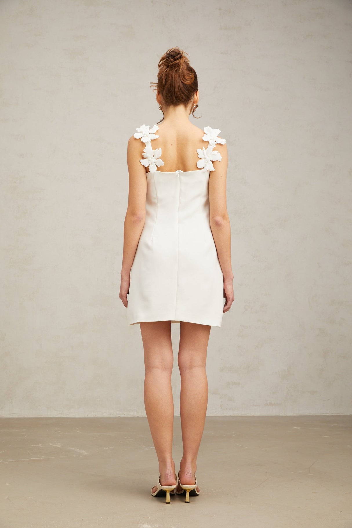 Young woman wearing a short white dress with floral embroidery standing in front of a plain background