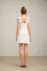 Young woman wearing a short white dress with floral embroidery standing in front of a plain background